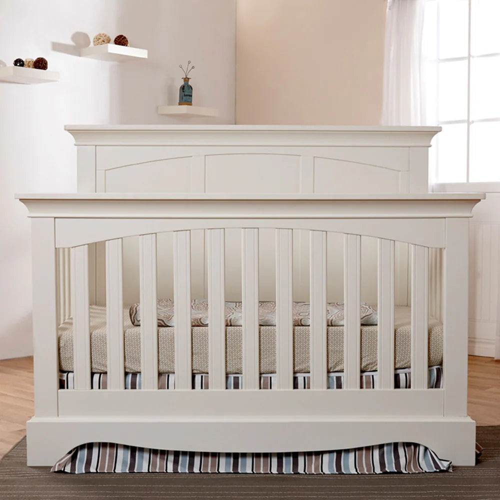 White crib with striped bedding in a room with a window and shelves.