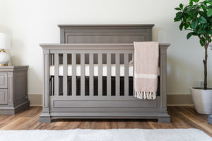 Ash crib with a white mattress and beige blanket in a room with a plant and nightstand.