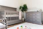 Load image into Gallery viewer, Nursery room with gray crib and dresser, white rug, and plant.

