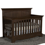Load image into Gallery viewer, A dark brown wooden convertible crib with a tall headboard and side rails, shown with a striped mattress and bedding on a gray shag rug against a white background.

