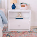 Load image into Gallery viewer, A white wooden nightstand with an open top shelf and a single drawer, styled in a children's bedroom setting with a blue lamp, books, and a basket on top.
