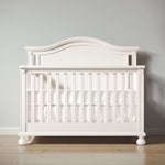 Load image into Gallery viewer, White crib with decorative headboard on a wooden floor against a gray wall
