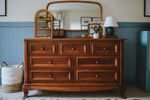 Load image into Gallery viewer, A warm brown wooden dresser with seven drawers and round knobs, styled in a bedroom setting with a matching wood-framed mirror, a small wicker birdcage decor piece, a lamp, and a woven basket on a blue paneled wall.
