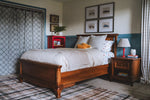 Load image into Gallery viewer, A medium brown wooden Antonio bed with a white comforter and patterned pillows, styled in a bedroom with blue wainscoting, a red side cabinet, and a patterned rug.
