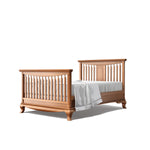 Load image into Gallery viewer, A wooden convertible crib bed with a warm medium-brown finish, shown with a white mattress and sheet on a solid white background.
