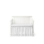 Load image into Gallery viewer, A white wooden crib, converted to a daybed, with a white mattress and ruffled crib skirt, shown against a solid white background.
