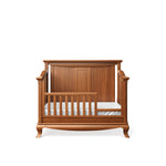 Load image into Gallery viewer, A wooden baby crib with a warm medium-brown finish, featuring a slatted front and side rails, a padded white mattress, and a matching wooden headboard panel, shown against a plain white background.
