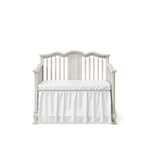 Load image into Gallery viewer, A white wooden convertible crib with a scalloped headboard, vertical slats, and a matching white mattress and ruffled skirt, shown on a plain white background.
