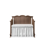 Load image into Gallery viewer, A wooden crib with a brown wood finish, featuring a white mattress and a white ruffled bed skirt, shown against a plain white background.
