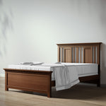 Load image into Gallery viewer, A wooden bed frame with a headboard and footboard in a warm medium brown finish, styled with white bedding and a white sheet, set against a minimalist light gray wall and light wood floor.
