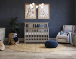 Load image into Gallery viewer, A styled nursery room featuring a light gray wooden crib as the central product, set on a round jute rug with a dark blue pouf. The room has a dark blue textured wall, a modern multi-light chandelier, two framed animal portraits, a cream recliner chair, and a potted green plant.
