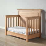 Load image into Gallery viewer, A light natural wood toddler bed with a tall headboard and side rails, shown with a white mattress on a light wood floor against a neutral gray wall.
