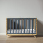 Load image into Gallery viewer, A modern wooden crib with a slate blue painted metal slat frame and natural wood sides and legs, shown with a white mattress against a neutral gray wall and light wood floor.
