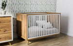 Load image into Gallery viewer, Wooden crib with white bedding in a room with a dresser and floral wallpaper.
