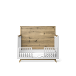 Load image into Gallery viewer, A modern baby crib, converted to toddler bed, with a white frame, white mattress, and a large natural wood headboard, shown against a plain white background.
