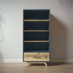 Load image into Gallery viewer, A tall, narrow bookcase with a dark blue painted frame and natural wood shelves and drawer front, shown standing on a light wood floor against a plain wall.
