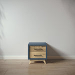 Load image into Gallery viewer, A small two-drawer nightstand with a slate blue painted frame and natural oak wood drawer fronts, shown against a plain light gray wall on a light wood floor.
