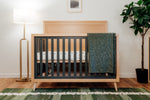 Load image into Gallery viewer, New York crib in natural and rosemary, shown in room setting with teal blanket, otted plane and gold floor lamp
