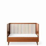 Load image into Gallery viewer, A mid-century modern wooden crib with a warm brown finish and vertical slats, shown with a white mattress on a clean white background.
