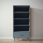 Load image into Gallery viewer, A tall, narrow bookcase with four open shelves and a single drawer at the bottom, finished in a muted slate blue-gray color with a small gold pull handle. It stands on tapered legs against a plain off-white wall and light wood floor.
