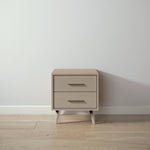 Load image into Gallery viewer, A two-drawer nightstand with a light beige wood finish and gold-toned metal handles, set on a light wood floor against a textured white wall.
