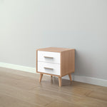 Load image into Gallery viewer, A small two-drawer nightstand with a light natural wood finish and white drawer fronts, shown in a minimalist room setting with a light gray wall and wood-look floor.
