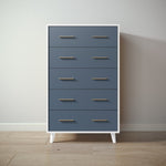 Load image into Gallery viewer, A five-drawer chest of drawers with a slate blue front and white frame, featuring slim gold metal handles and white legs, set against a neutral wall and light wood floor.
