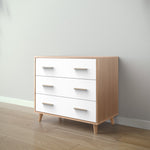 Load image into Gallery viewer, A three-drawer chest of drawers with a light natural wood frame and white drawer fronts, shown in a minimalist room setting with light wood flooring and a gray wall.
