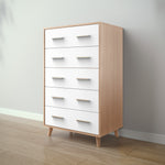 Load image into Gallery viewer, A five-drawer chest of drawers with white fronts and light natural wood sides, legs, and handles, shown in a minimalist room setting with a gray wall and light wood floor.
