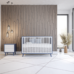 Load image into Gallery viewer, A modern nursery setup featuring a white crib with blue trim and matching blue legs, a small white and blue nightstand, and a large textured wood accent wall behind it. The room has white tiled flooring, pendant lights, and a potted palm near a window.

