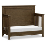 Load image into Gallery viewer, A dark brown wooden crib converted to a daybed with a white mattress, shown from a three-quarter angle on a white background.
