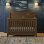 Load image into Gallery viewer, A wooden crib with a dark brown finish stands in a styled nursery setting with a blue paneled wall, two gold wall sconces, and a patterned rug.
