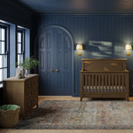 Load image into Gallery viewer, A styled nursery room with dark blue paneled walls and ceiling, featuring a wooden crib, a wooden dresser, an arched door, and a patterned area rug.
